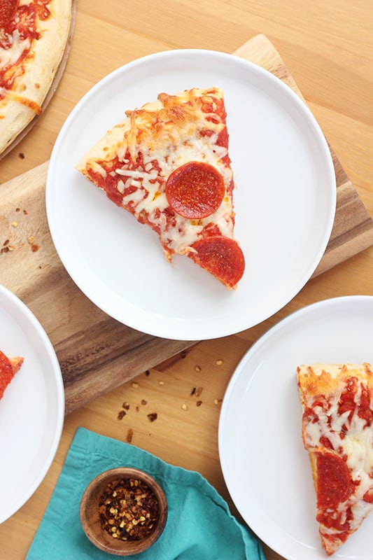 three slices of pizza sitting on white round plates on a wooden tabletop with red pepper flakes sprinkled around