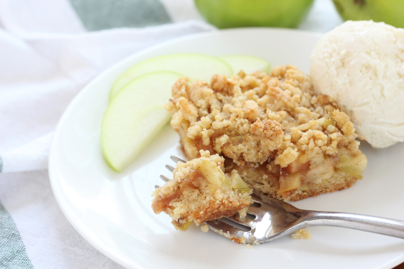 apple crumble square sitting on a small with plate with a bite sitting on a fork with green apple slices sitting off to the side with a scoop of vanilla ice cream