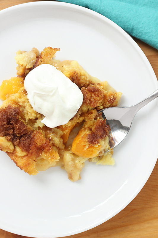 top down image of a white round plate with a serving of a peach cobbler sitting with a bite on a spoon and a dollop of whipped topping in the center