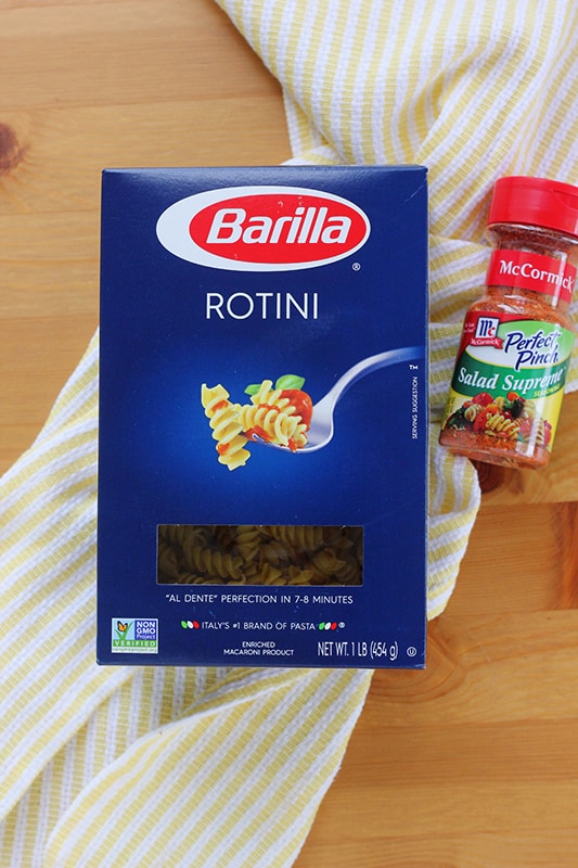 blue box of Barilla rotini pasta and a container of McCormick salad supreme seasoning sitting on a white and yellow striped napkin on a wooden table top