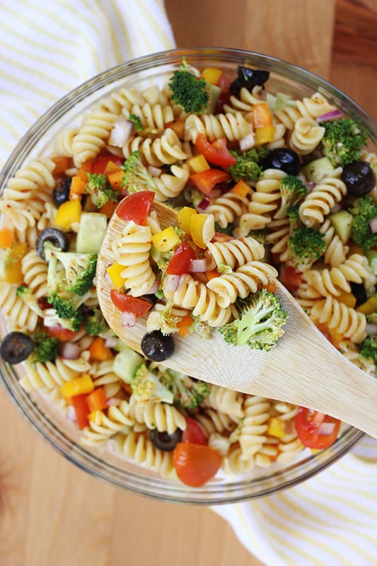 top down image of pasta and vegetables in a glass mixing bowl with some of the salad on a wooden spoon that is hovering over the bowl