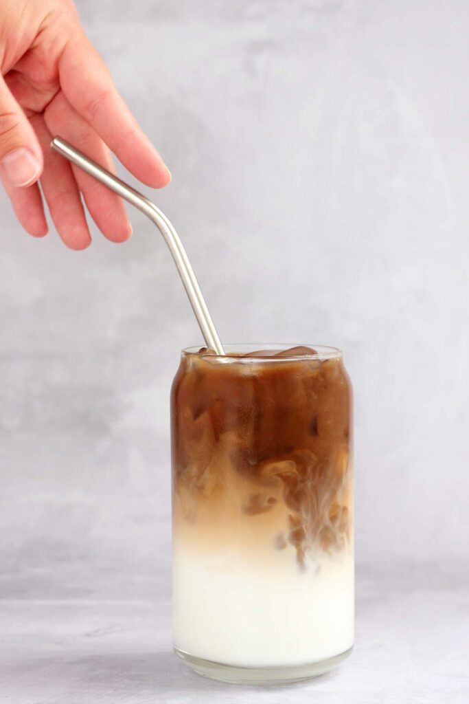 Vanilla Latte on One Sweet Appetite hand reaching for a metal spoon that is sitting in a glass with ice, milk, and coffee