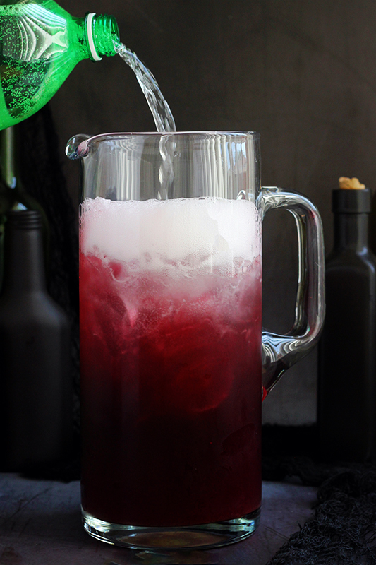 tall thinner pitcher filled with a purple drink with a lemon lime soda being poured into the top of the pitcher causing it to foam. 