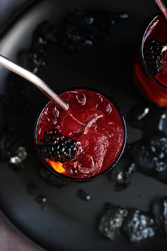 Top down image showing two skinny glasses filled with a purple drink with blackberries and orange slices floating among the ice. Glasses are sitting on a black plate with melting ice pieces