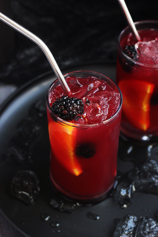 two skinny glasses filled with a purple drink with blackberries and orange slices floating among the ice. Glasses are sitting on a black plate with melting ice pieces