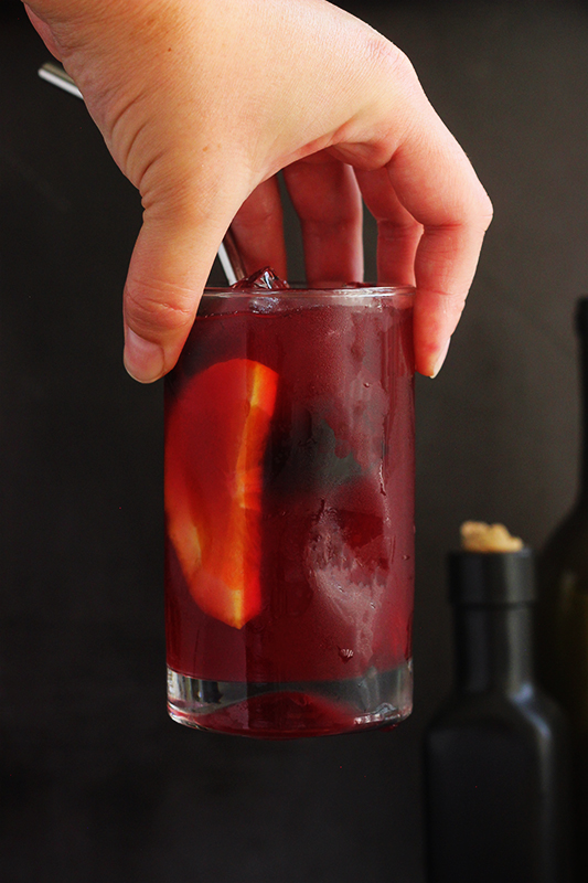 hand holding a slightly thinner glasss that is filled with a purple drink, blackberries, and an orange slice in front of a black background