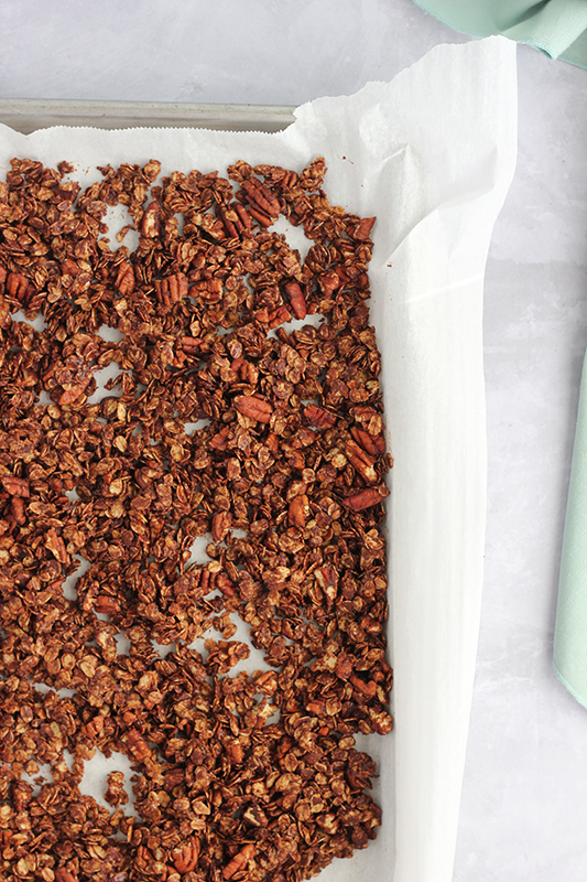 chocolate granola baked and spread on a sheet pan that has been lined with parchment paper