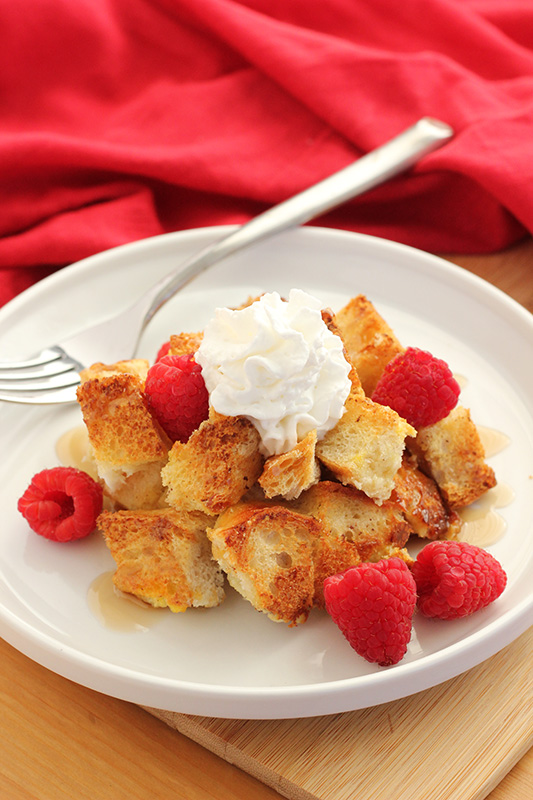 french toast bake on a white plate topped with syrup, raspberries, and whipped cream