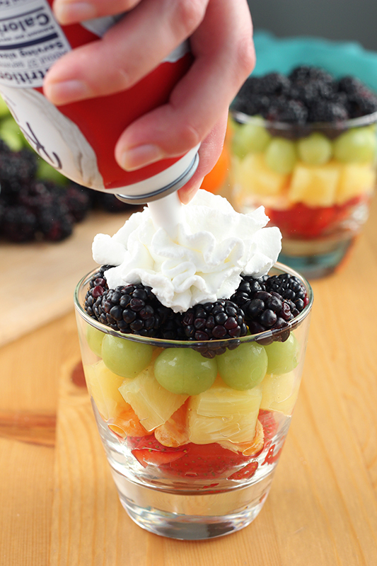whipped cream being sprayed onto the top of a cup of layered fruit