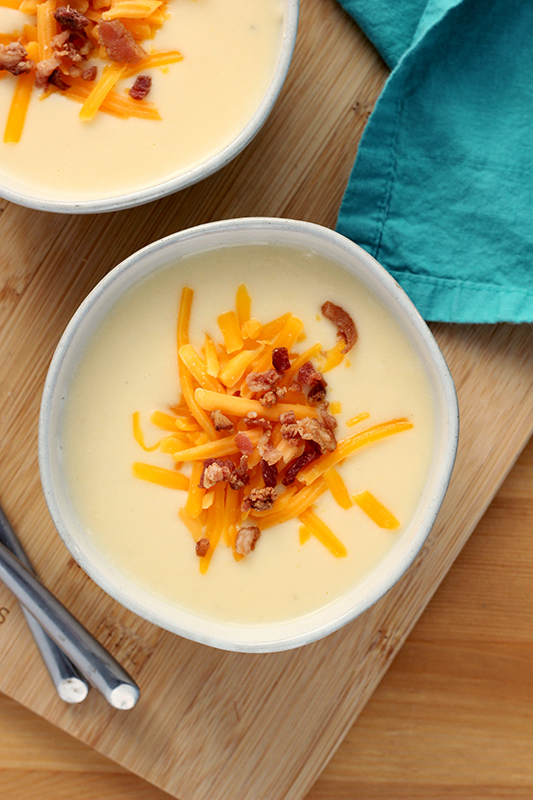 two bowls of cheese soup topped with shredded cheese and bacon on a wooden cutting board