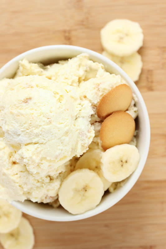 top down image of a white bowl sitting on a wooden tabletop with scoops of banana ice cream, slices of bananas, and vanilla wafer cookies