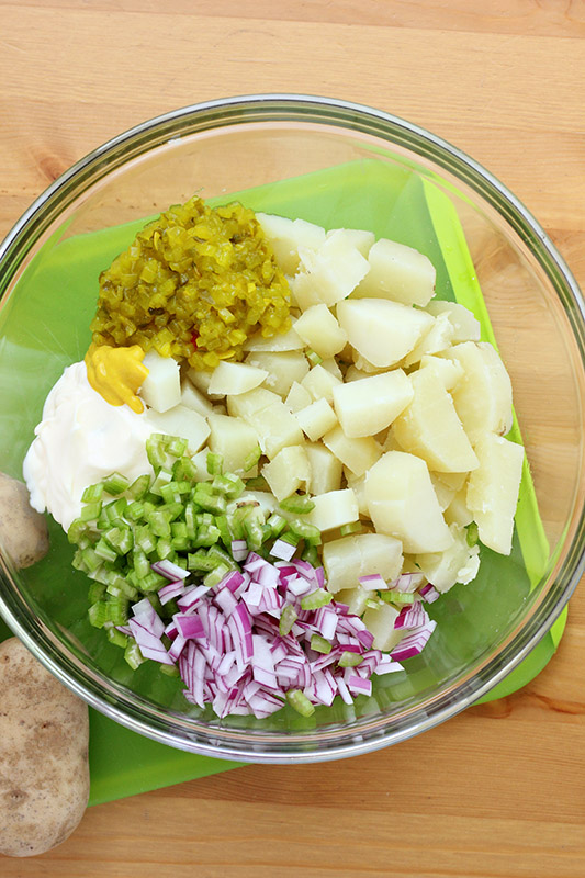 peeled and cut cooked potatoes, relish, mayo, celery and red onion in a glass mixing bowl sitting on a green cutting board