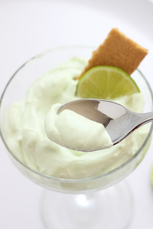 tall martini glass filled with a green mousse that has a bite being scooped with a silver spoon. A graham cracker rectangle and lime wedge sitting behind the spoon