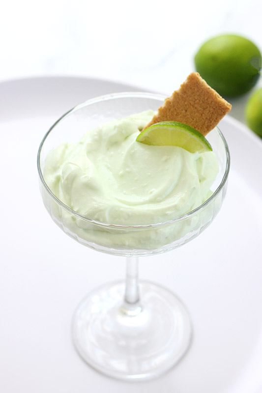 tall martini glass filled with a green mousse that has a bite being scooped with a silver spoon. A graham cracker rectangle and lime wedge sitting behind the spoon