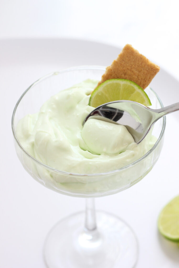 tall martini glass filled with a green mousse that has a bite being scooped with a silver spoon. A graham cracker rectangle and lime wedge sitting behind the spoon