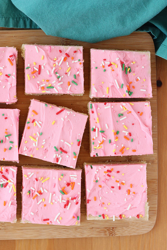 sugar cookie bars topped with pink frosting and colored sprinkles on a wooden cutting board