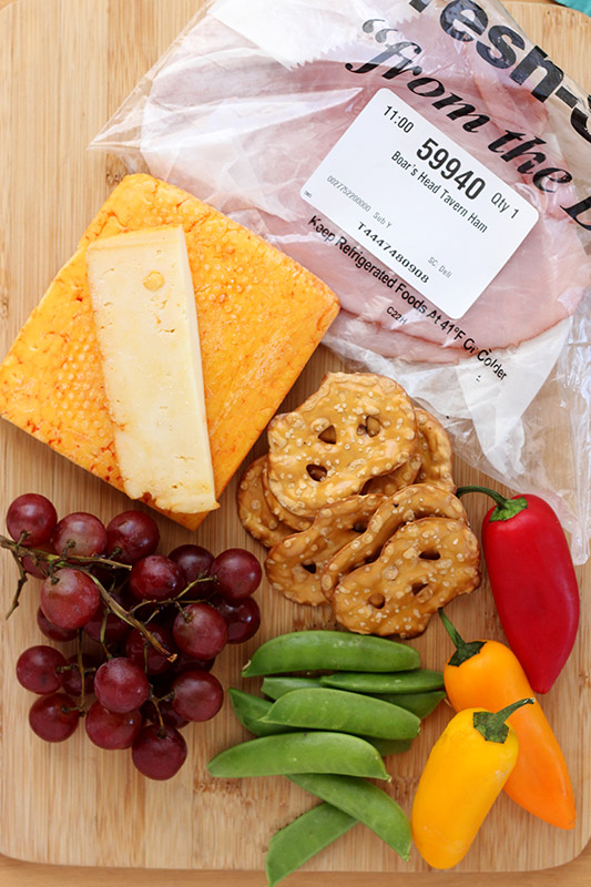 ham, cheese, pretzels, grapes, peas, and peppers on a wooden cutting board