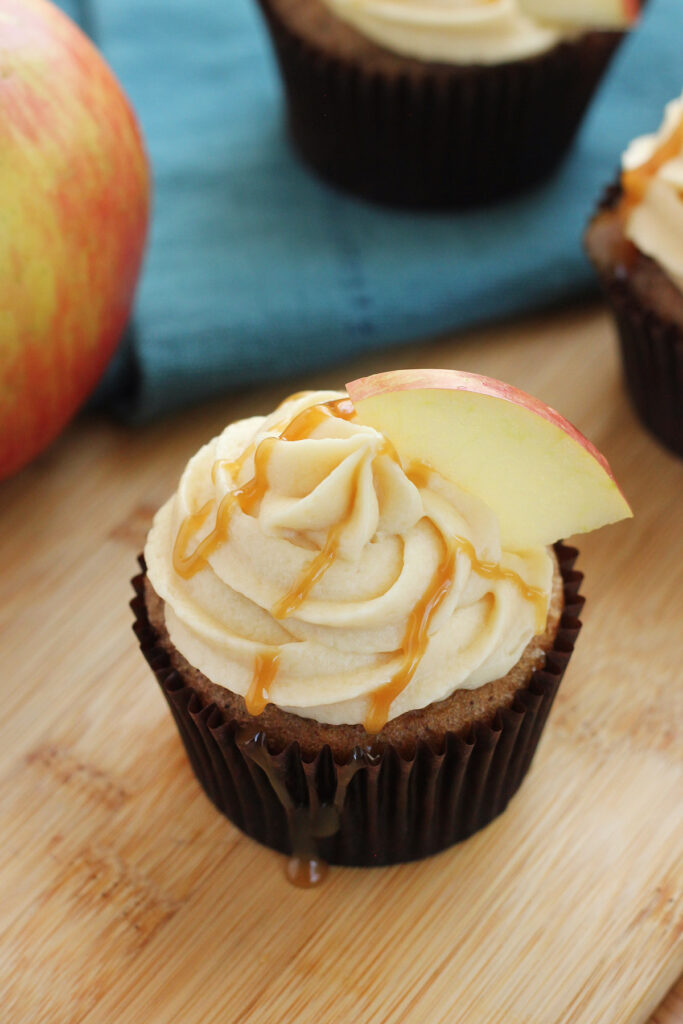 caramel drizzled cupcake sitting on a wooden cutting board with an apple slice on top. 