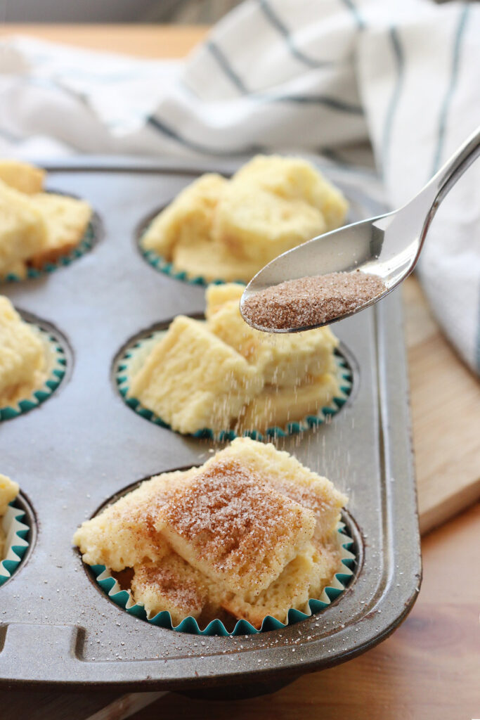 spoon sprinkling cinnamon and sugar over top of bread cubes inside a muffin tin