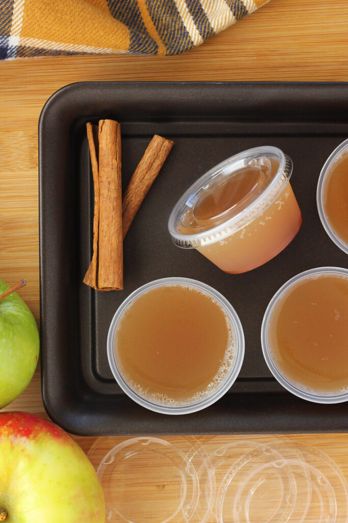 plastic containers filled with apple jello shots with cinnamon sticks and apples on the side.