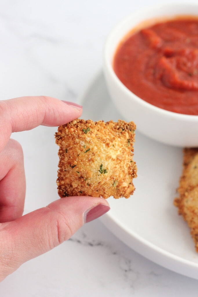 single toasted ravioli being held over a bowl of sauce