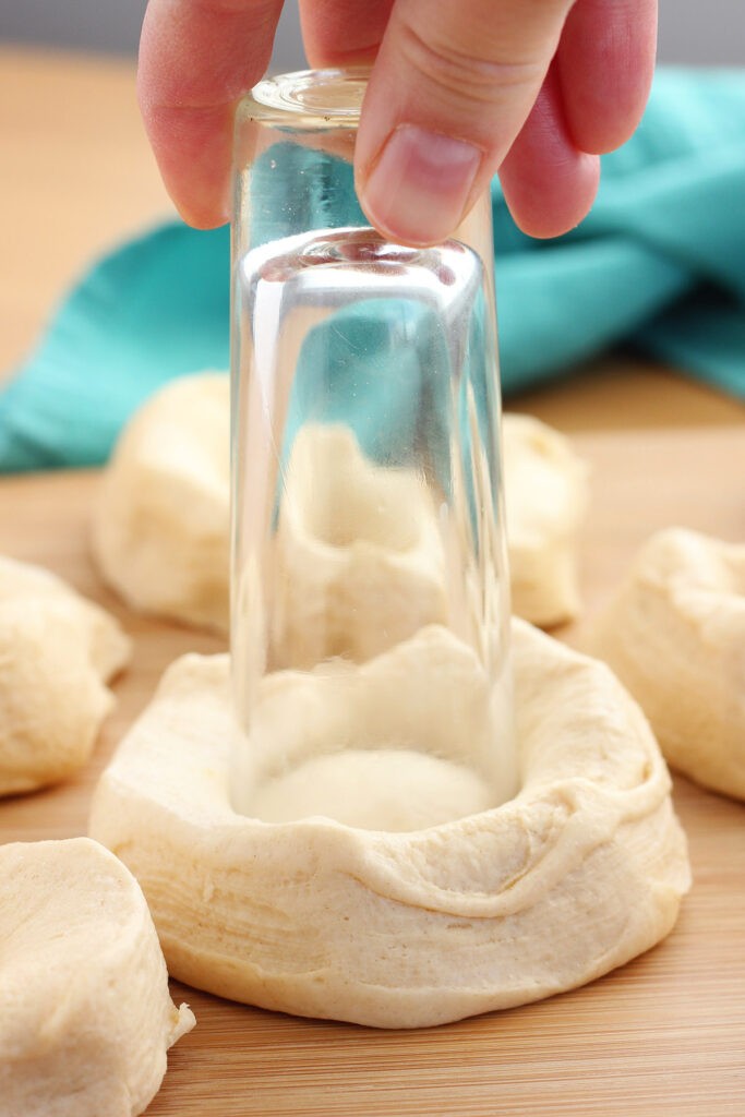biscuit dough sitting on a wooden cutting board with the center being cut out by a tall shot glass