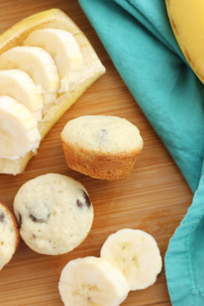 mini banana pancake muffins ona wooden cutting board with sliced bananas and a teal napkin