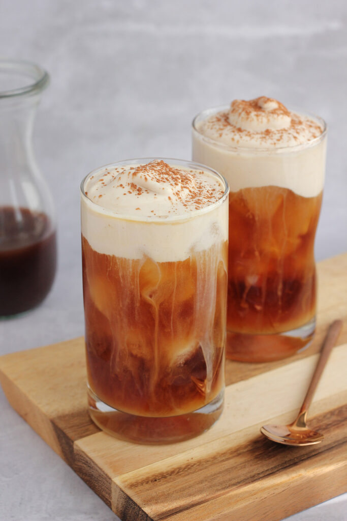 two tall classes sitting on  a dark wood rectangle serving board filled with cold brew and topped with foam that has been dusted with cocoa powder