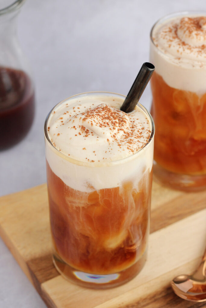 close up image of a glass of iced coffee that is topped with a layer of slightly under whipped cream, sprinkled with cocoa powder, and a black straw
