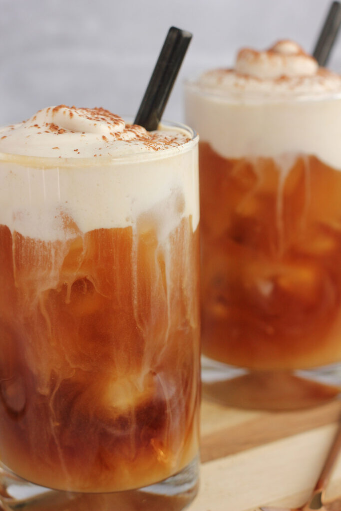 close up image of two iced coffees with a swirl of cream falling into the glass. Topped with foam and a black straw