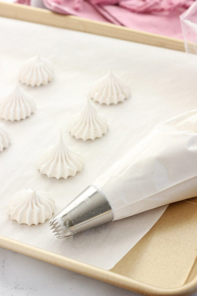 piping bag fitted with a star attachment and filled with meringue on a baking tray that is lined with parchment paper with small dots of meringue piped onto the tray