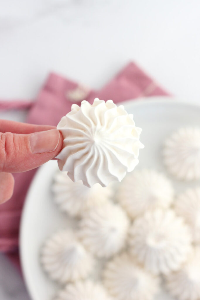 hand holding a single white cookie made from whipped egg whites in a spiral shape over a white plate that is topped with more cookies and a pink napkin