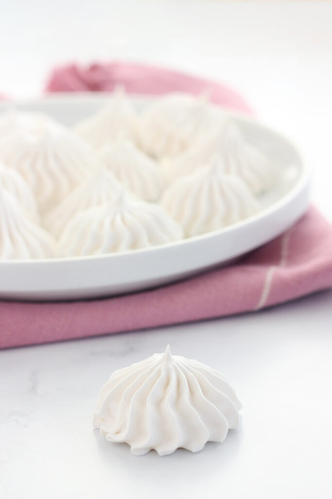 single meringue cookie sitting on a white table with a plate of cookies in the background sitting on a pink napkin.