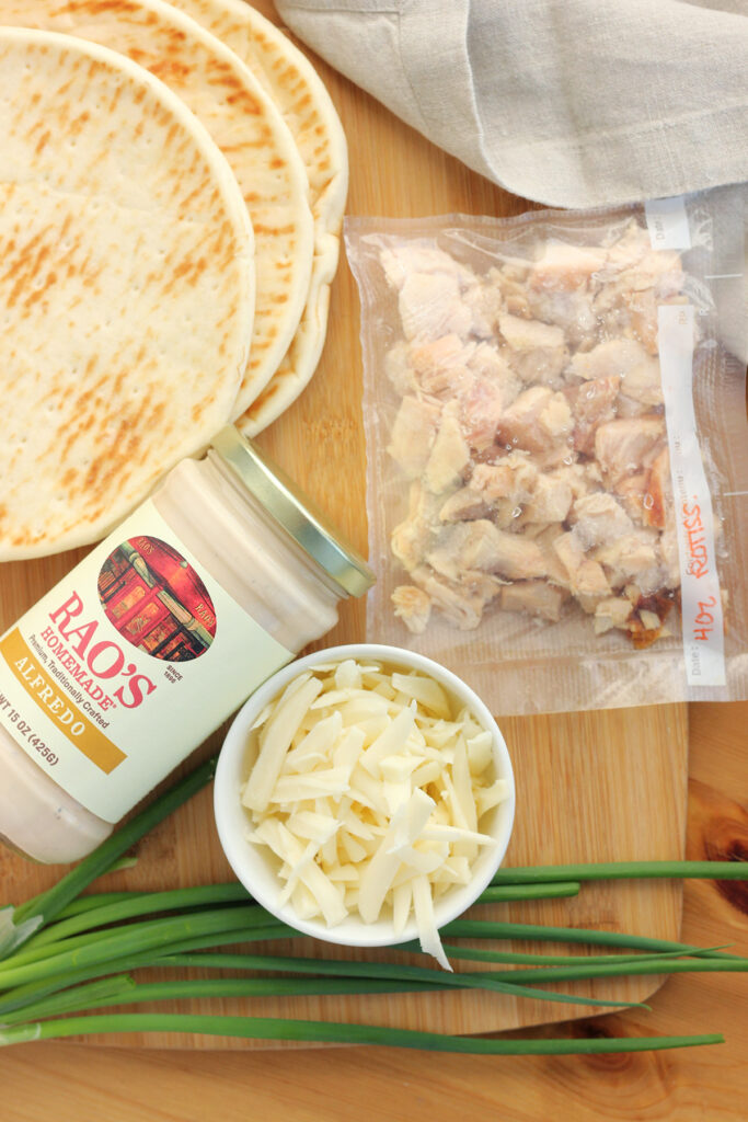 wooden cutting board topped with flatbread, jar of alfredo sauce, bag of rotisserie chicken, white bowl of shredded mozzarella cheese and fresh green onions