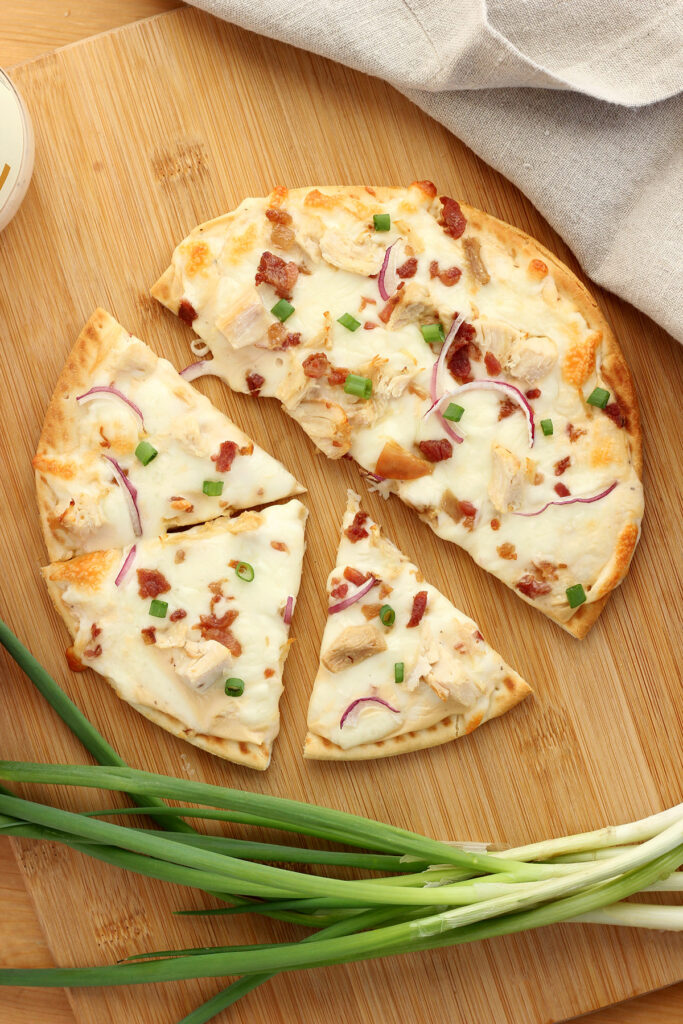 flatbread chicken pizza sitting on a wooden cutting board with half of it cut into 3 triangles. Tan napkin and green onion sitting off to the side