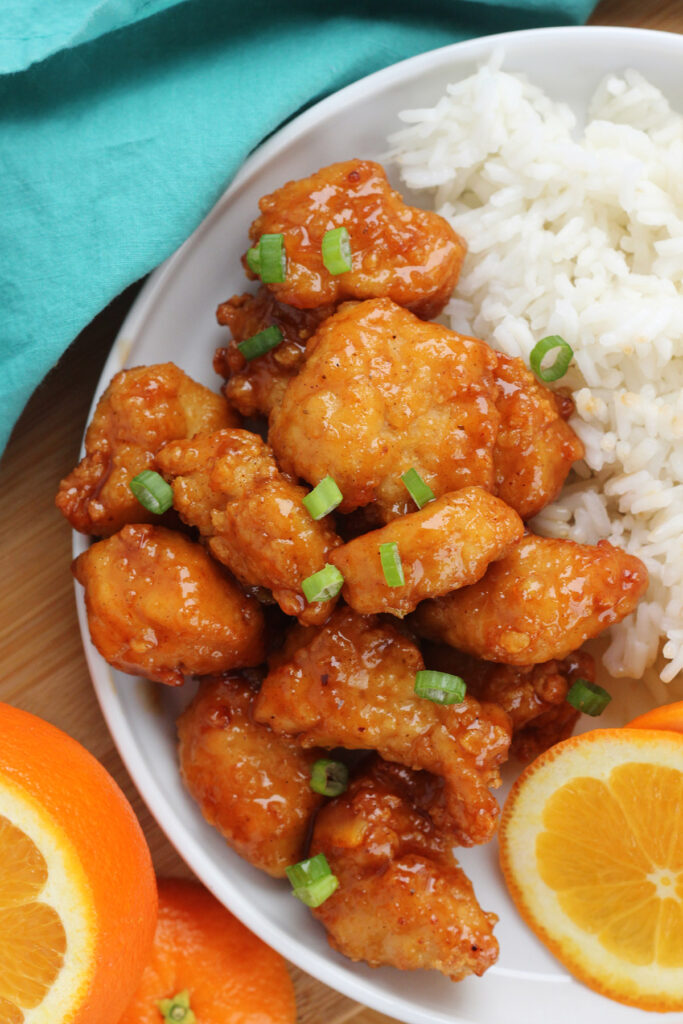 Stack of orange chicken on a white plate with a side of rice and orange slices. All sitting on a wooden cutting board with a teal napkin and more orange slices