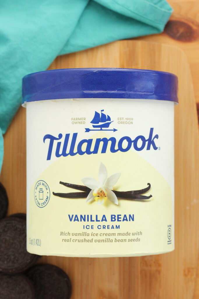 container of Tillamook vanilla bean ice cream laying on top of a wooden cutting board with chocolate sandwich cookies sitting off to the side with a teal napkin