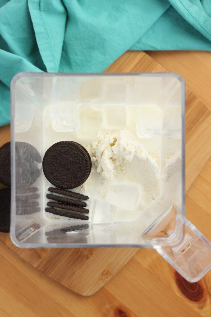 top down image of a blender without a lid on sitting on a wooden cutting board. Inside is milk, ice cream, ice and chocolate sandwich cookies