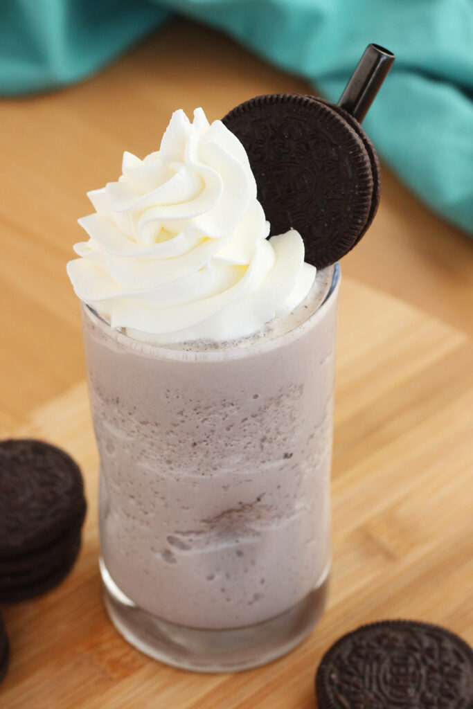 glass filled with a chocolate milkshake topped with whipped cream and a single oreo cookie with a dark black straw. All sitting on a wooden cutting board with additional oreo cookies and a teal napkin