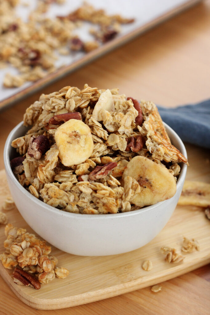 off white bowl sitting on a small wooden cutting board filled with granola that includes pecans and dried bananas