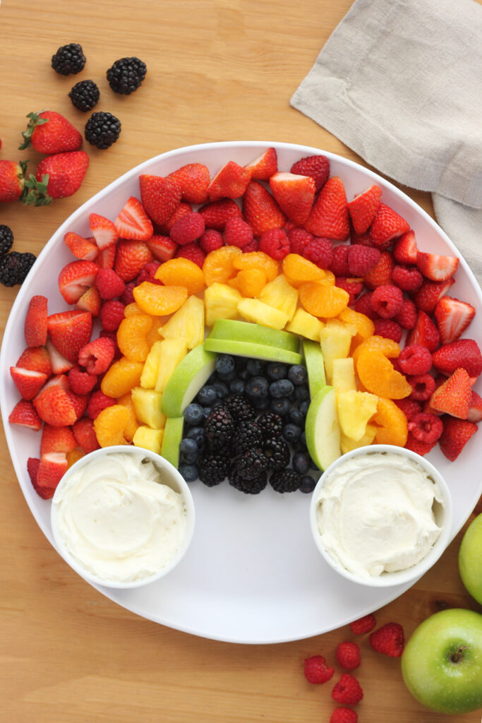 white round serving tray sitting on a wooden table with a colorful rainbow display of fruit and two small white bowls of fruit dip
