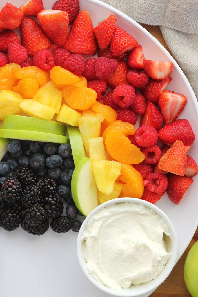 close up image of a fruit platter that is layered in a rainbow color scheme on a white round serving board with a white round bowl that is filled with fruit dip