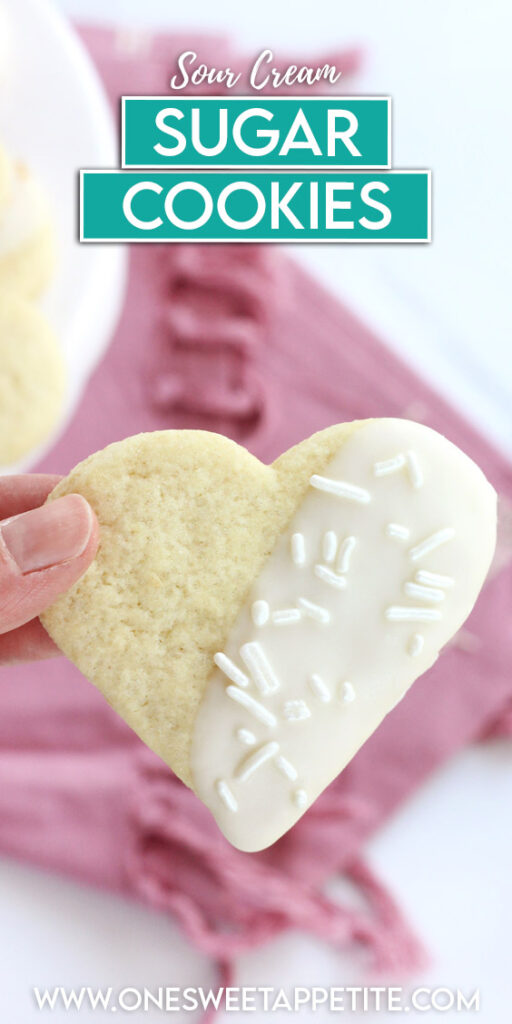 pinterest graphic of a heart shaped cookie that has been half dipped in chocolate held over a pink napkin with text overlay reading "sour cream sugar cookies"