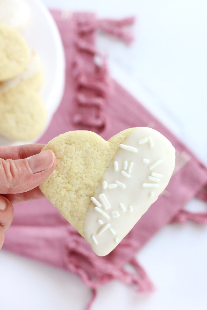 heart shaped cookie that has been half dipped in white chocolate and sprinkled with white jimmies being held over a pink napkin