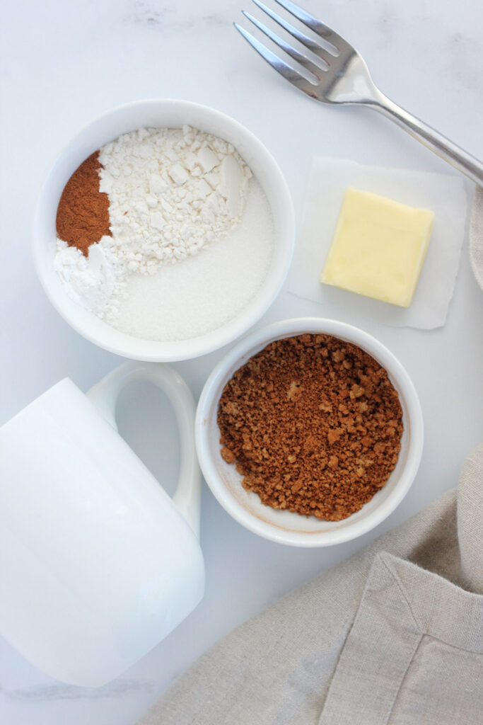 white mug laying sideways on a white table top with two bowls filled with mug cake ingredients and a tab of butter sitting off to the side. 