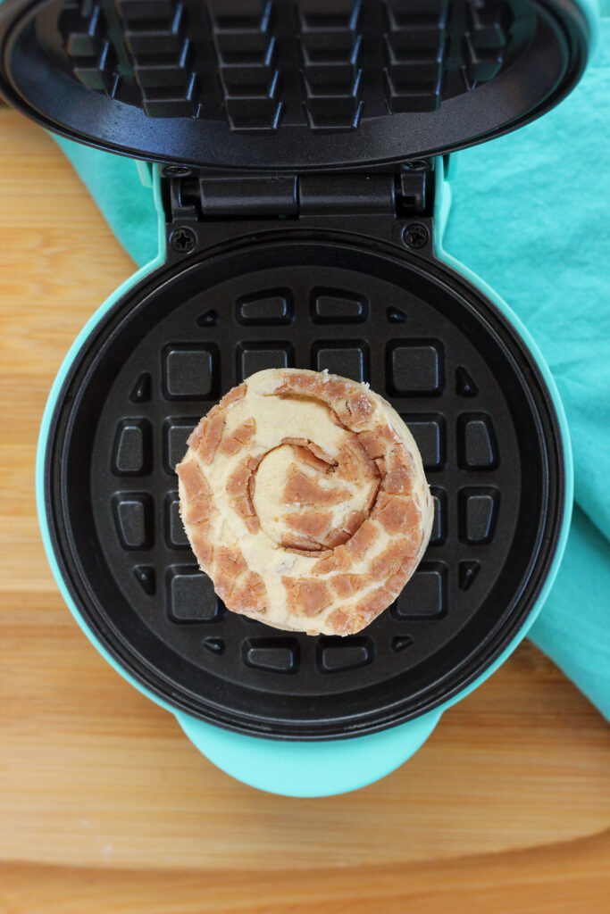 open waffle maker with a single unbaked cinnamon roll sitting in the center on top of a wooden table and teal napkin