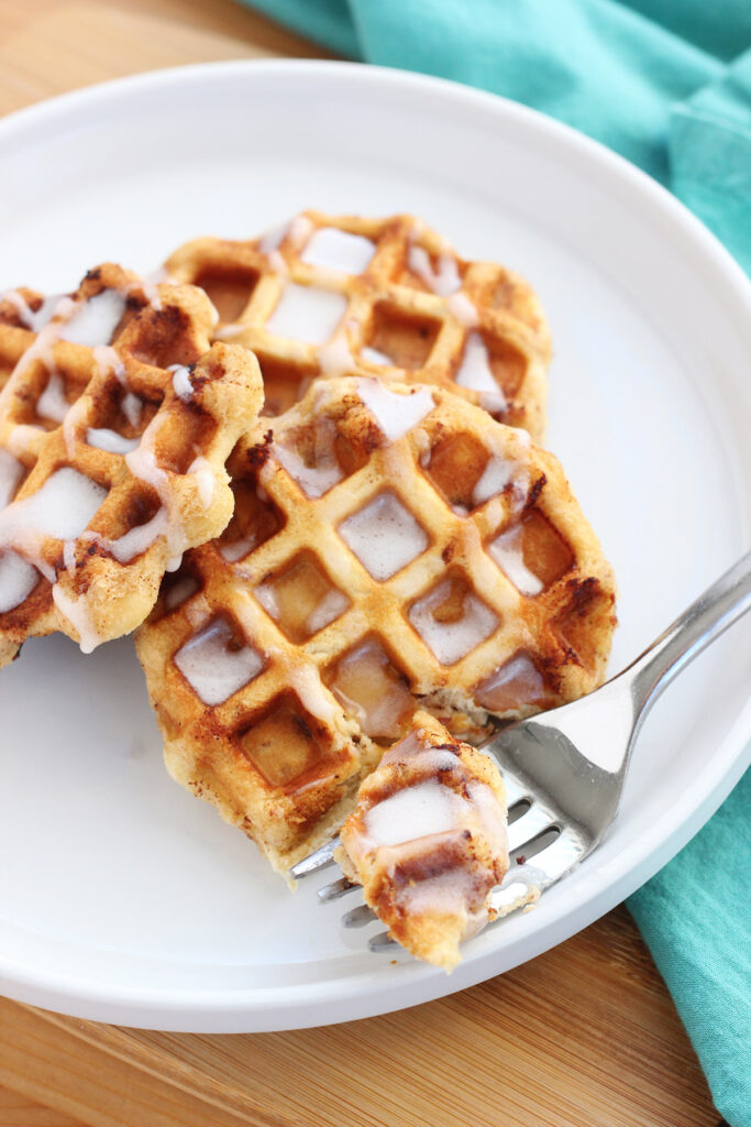close up image of a white plate with three cinnamon rolls that have been cooked in a waffle iron. Drizzled with glaze with one bite sitting on a fork. 