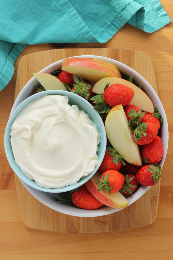 large white bowl filled with fresh strawberries and apple slices with a smaller blue bowl sitting off to the side filled with sweet dip