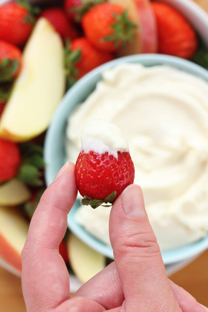 top down image of a blue bowl filled with dip and a spread of apples and strawberries. Single strawberry that has been dipped in the dip is being held over the top of the bowl