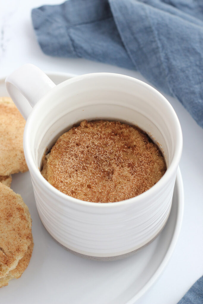 white mug sitting on a white plate with cookies on the side. Inside is filled with a cake that is topped with cinnamon and sugar